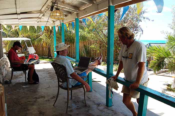 Two men sit in beach chairs on laptop computers under a wooden structure. A third man stands with his hands on the railing to the structure.