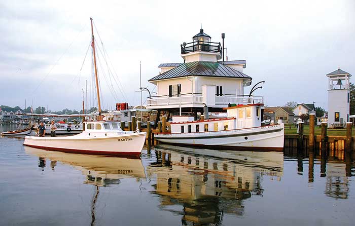 Two white wooden boats sit outside of the Chesapeake Bay Maritime Museum, a nautical white building resembling a lighthouse.