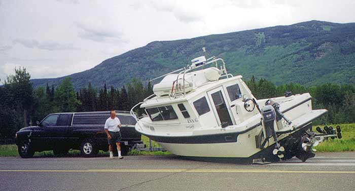 A large boat tips over into the road, apparently fell off the trailer it was on. A man steps out of his black SUV to look at the boat.