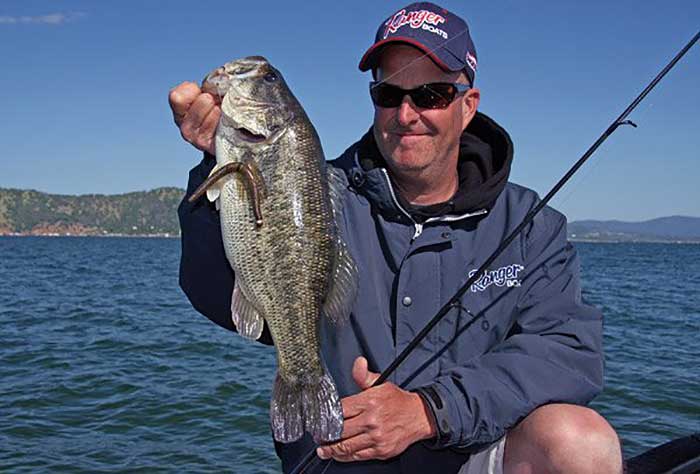 Man wearing sunglasses, a ball cap and a jacket stands holding fishing rod and large bass catch