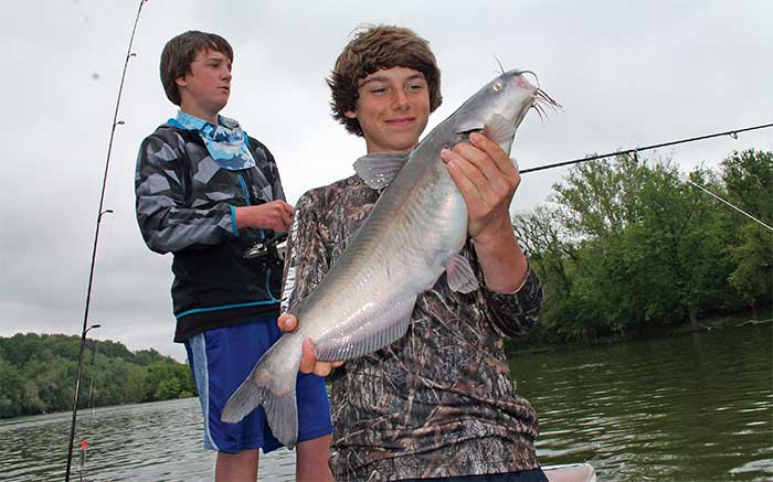 Two boys standing in a boat on a water one holding a blue catfish he just caught with fishing rods visible in the background