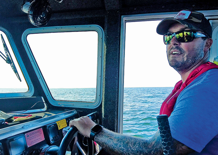 Adult male in wearing a baseball hat, black sun glasses and red life jacket in the cockpit of a vessel.