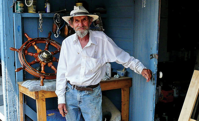 Elderly man wearing a large white hat, white button down shirt and blue jeans posing for a photo in front of a wooden shed.