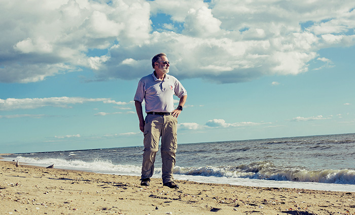 Elderly adult male wearing a polo and khaki pants standing on the beach during an overcast day.