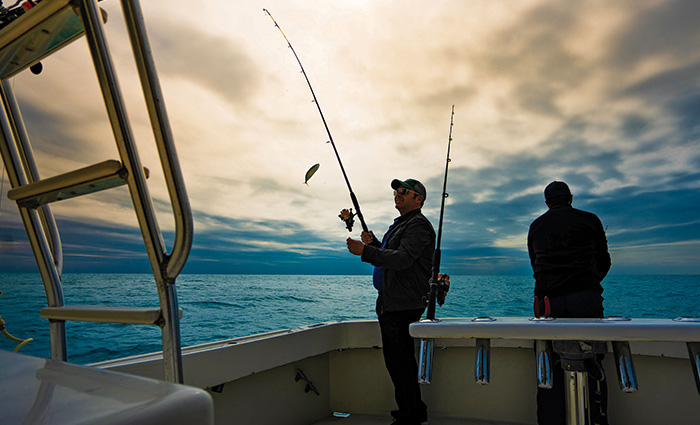 Two men fishing on the back of a white boat as the sun sets on open waters.