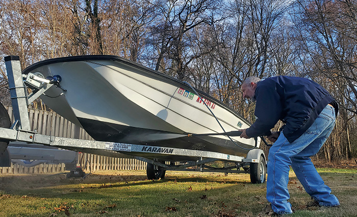Elderly adult male wearing jeans and a blue jacket crouching to power wash a white fishing boat.