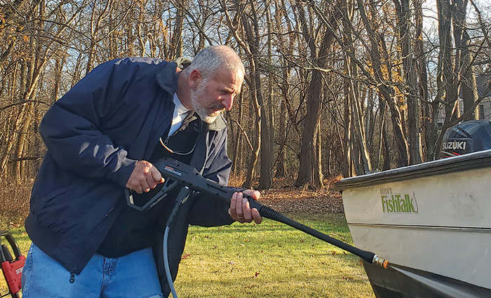 Senior adult male with a white beard wearing a navy jacket and jeans leaning into power wash a white and black boat.