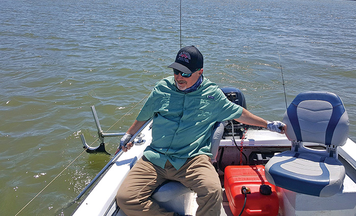 Senior male wearing a black ballcap, sunglasses, blue shirt and brown pants using a commercially made stick as a trotline on a fishing boat.