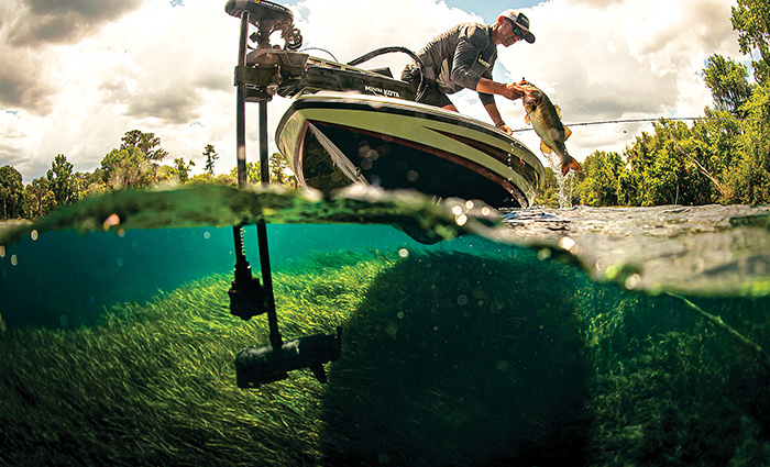 View from below of an adult male wearing long-sleeve gray shirt and ballcap catching a large bass