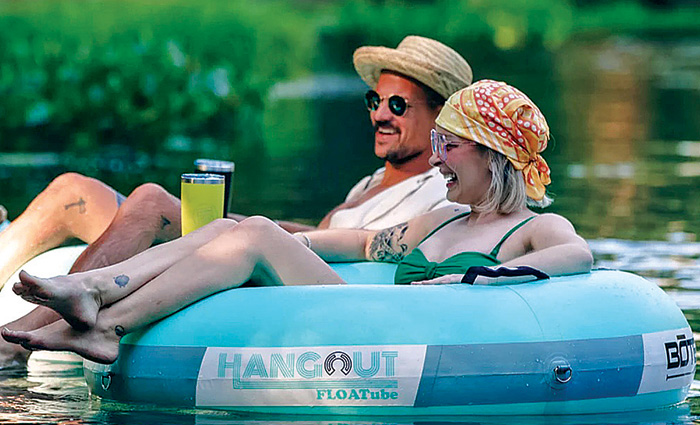 Two adults wearing sunglasses and enjoying beverages while lounging on light blue tubes on the water.
