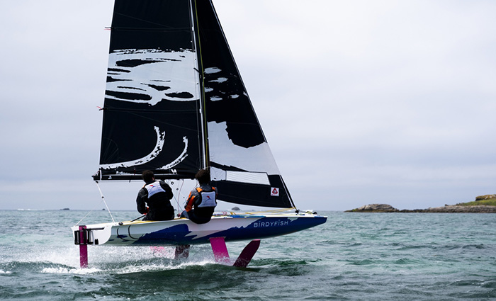 Two adults with white lifejackets on open waters aboard a blue and white sailboat with black and white sails.