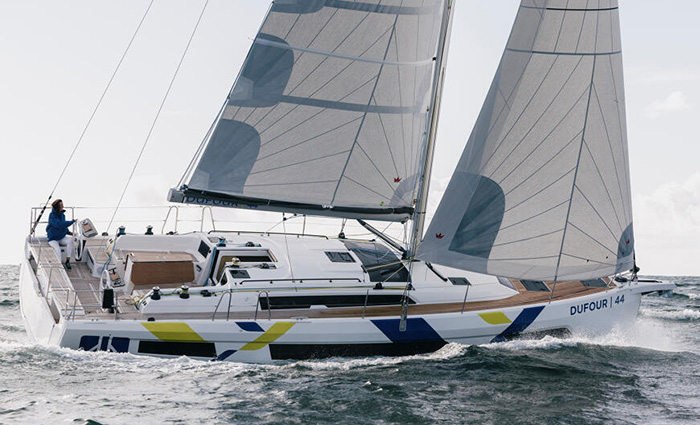 Adult wearing white pants and a blue jacket navigating a large white with yellow and black striped sailboat out at sea.