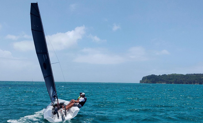 Two adults sailing aboard a white sailboat with navy sails on a sunny day with land in the background.