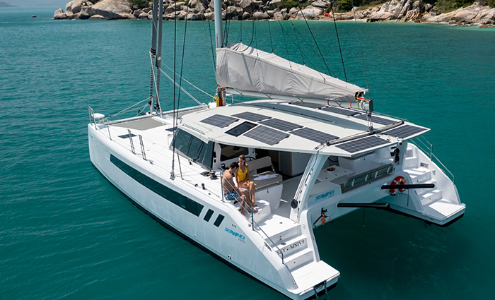 An adult male wearing red and blue swim trunks sitting beside an adult female in a yellow swimsuit aboard a large white sailboat with solar panels on the water.