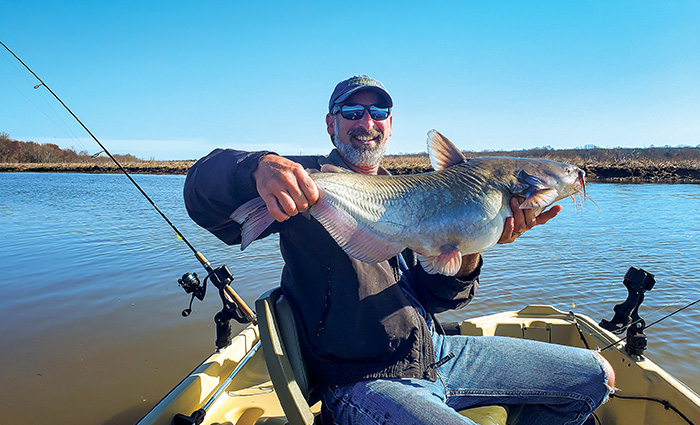 Bearded adult male wearing a green ballcap, sunglasses, jeans and a black jacket proudly displaying a fish caught on a small boat.