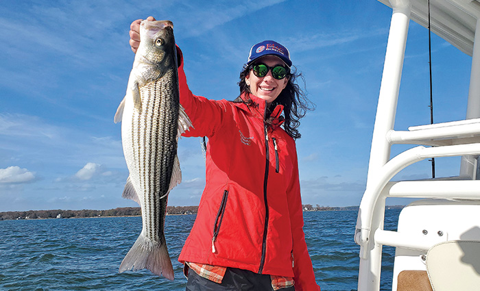 Brunette female wearing a ballcap, black sunglasses and a red jacket proudly displaying a caught fish. 