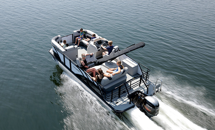 Arial view of three children and four adults aboard a white pontoon boat speeding across open waters.
