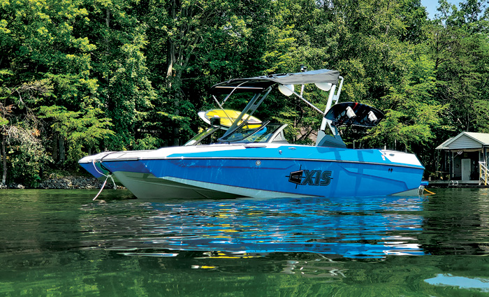 Blue and white  boat with two yellow wakeboards attached anchored on a lake with lush green trees in the background.