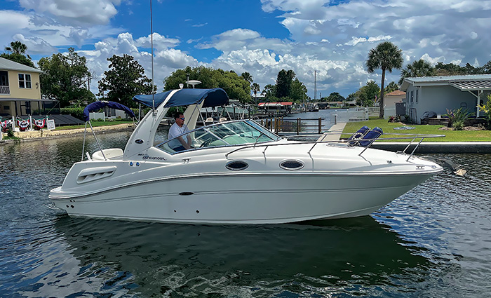 Adult male wearing a white polo and sunglasses steering a large white boat out of a dock on an overcast day.