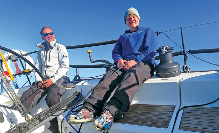 An adult male wearing a white jacket and black sunglasses sitting beside a young adult wearing a knit hat, blue sweatshirt and black pants aboard a white boat.