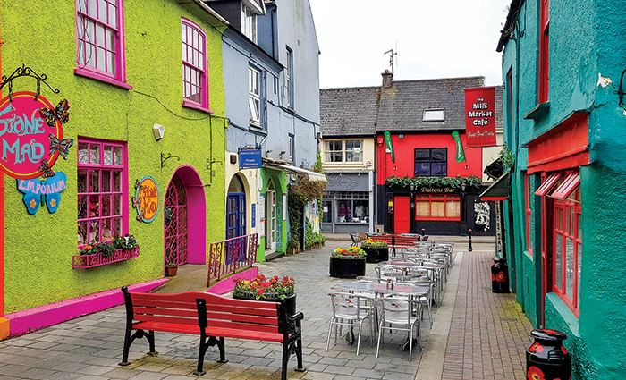 Colorful side streetscape with green, teel and blue buildings, large flower pots, white chairs and tables and a large red bench. 
