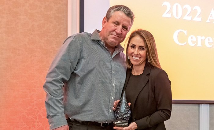 Man  in a gray button up shirt posing for a photo next to a brunette female in a black suit holding an award.