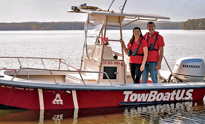 An adult male and adult female wearing red polo shirts and red life jackets aboard a small red boat out on a lake.
