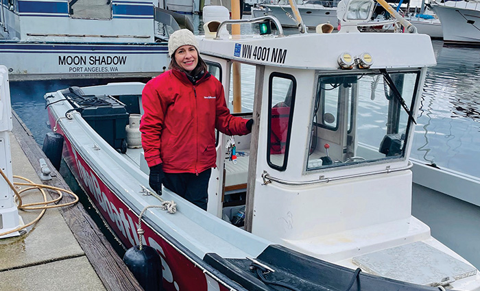 An adult female wearing a white knit hat, red jacket and black gloves posing for a photo aboard a small boat on a dock.