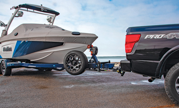 A white and blue boat with a gray cover on a blue trailer being pulled by a black truck along a beach.