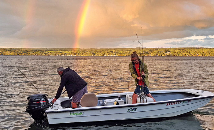 An adult male wearing gray shorts and a black hooded jacket and an adult female wearing a red shirt and tan jacket aboard a small white fishing boat with a rainbow in the background.