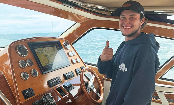 Young adult wearing a hooded sweatshirt and hat giving a thumbs up at the steering wheel of a boat.