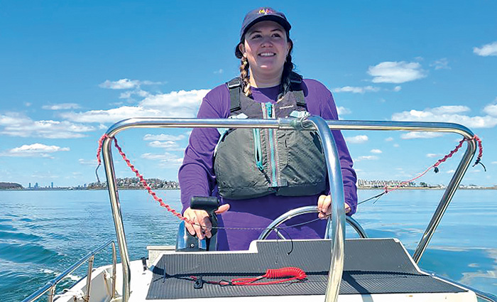 Young adult female wearing a black life jacket and red long sleeve shirt steering a white boat on open waters.