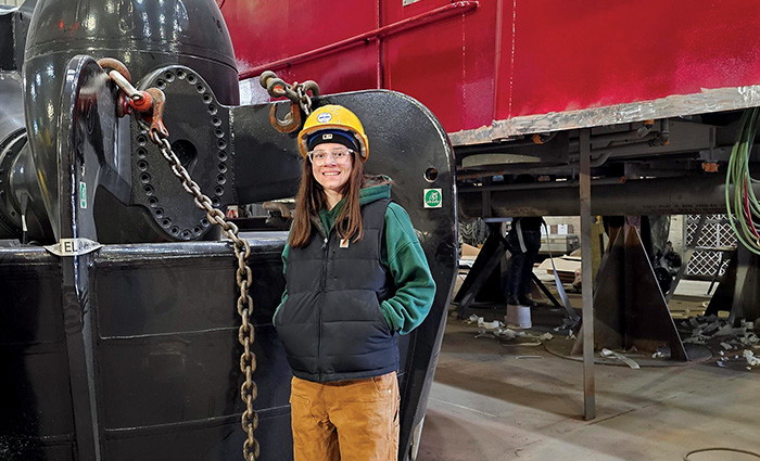 Young adult female wearing a black vest, green hooded sweatshirt, brown pants and a yellow hard hat posing for a photo.