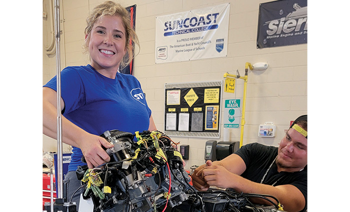 Blonde young adult female wearing a blue shirt smiling as she holds up a piece of machinery.