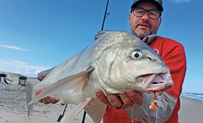 Adult male wearing a ballcap, black rimmed glasses and a red jacket proudly displaying a large fish caught.