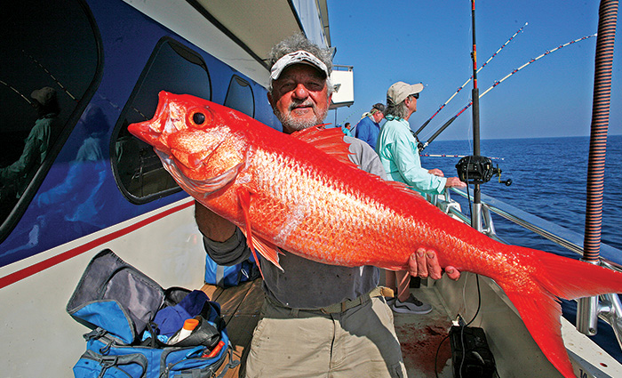 Adult male with a beard wearing a white visor, gray shirt and tan pants holding a large bright orange fish.