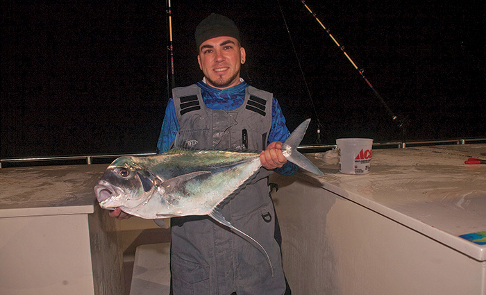 Young adult male wearing blue shirt and gray overalls holding a large fish on a boat at night.