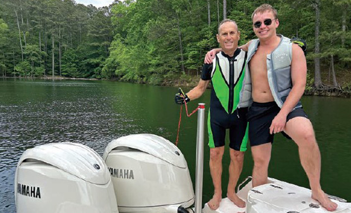 Tow adult males wearing lifejackets standing at the stern of a boat with dual motors on the water.