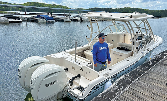 Man wearing a ballcap, sunglasses and long-sleeve blue shirt standing on a white vessel next to a dock.