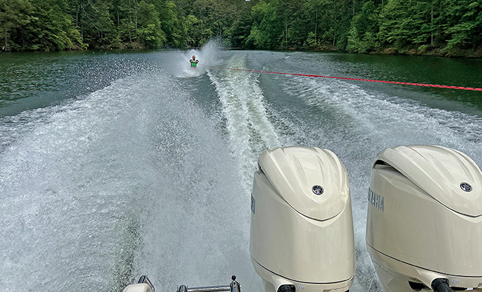 View from the stern of a boat of a man wakeboarding with lush green trees and water in the background.