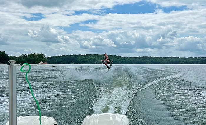 Outward view from the stern of a boat showing a young adult airborne while wakeboarding. 