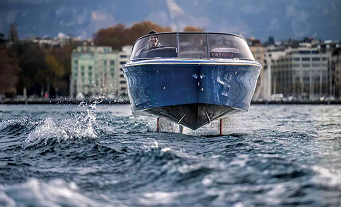Front view of an adult male driving a blue hydrolift powerboat on the water with various buildings in the background.