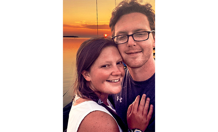 Young adult female wearing a white dress and young adult male with glasses taking a selfie with a sunset in the background.