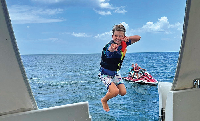 Young boy wearing a multi-colored life jacket pointing while jumping off a boat into clear water.