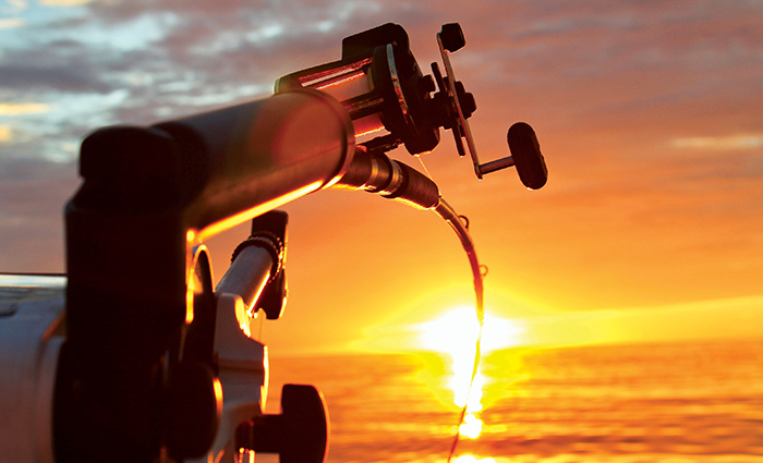 A fishing reel attached to a boat bending while the sunsets against the ocean at sunset.
