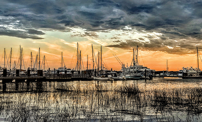 Numerous sailboats docked on the water as the sun peaks out of dark clouds.
