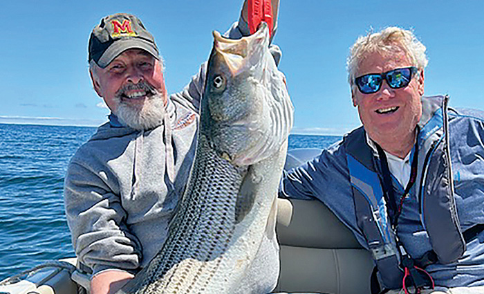 Two senior adult males proudly showing off a large base fish while on a boat in open waters.