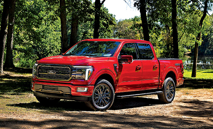 Red four door Ford truck parked amongst trees on a sunny day.