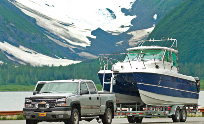 A silver four door Chevrolet pickup truck parked alongside a road with a large blue and white boat on its trailer and snow covered hills in the background.