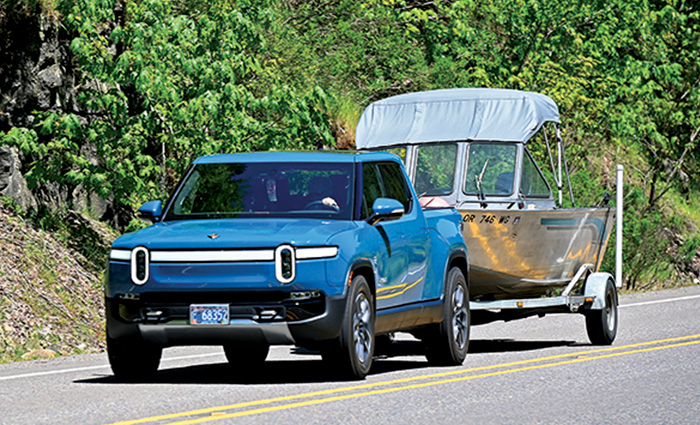 Bright blue four door truck towing a small silver boat along a two lane road on a sunny day.